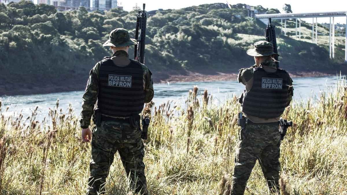 Polícia de Fronteira: Dois policiais militares do BPFron, vestidos com uniforme camuflado e colete, observam a margem de um rio armados, com área de vegetação e ponte ao fundo.