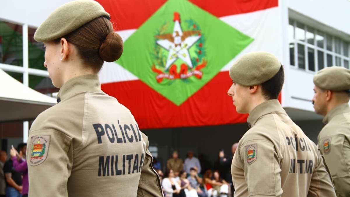 soldado e bombeiro temporário: Imagem mostra policiais militares de Santa Catarina, uniformizados e perfilados, com a bandeira do estado ao fundo.