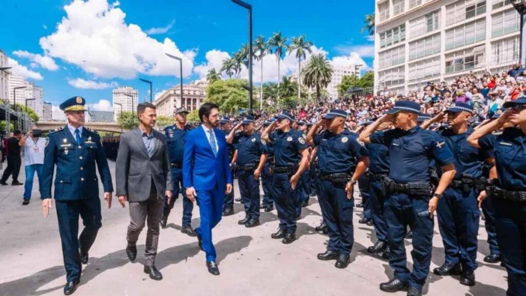 Formação de guardas municipais em São Paulo com presença de autoridades. Agentes uniformizados prestam continência enquanto desfilam diante de gestores da segurança pública, em evento ao ar livre com grande público nas arquibancadas. A imagem simboliza a valorização e militarização da Guarda Civil sob nova identidade de "Polícia Municipal".