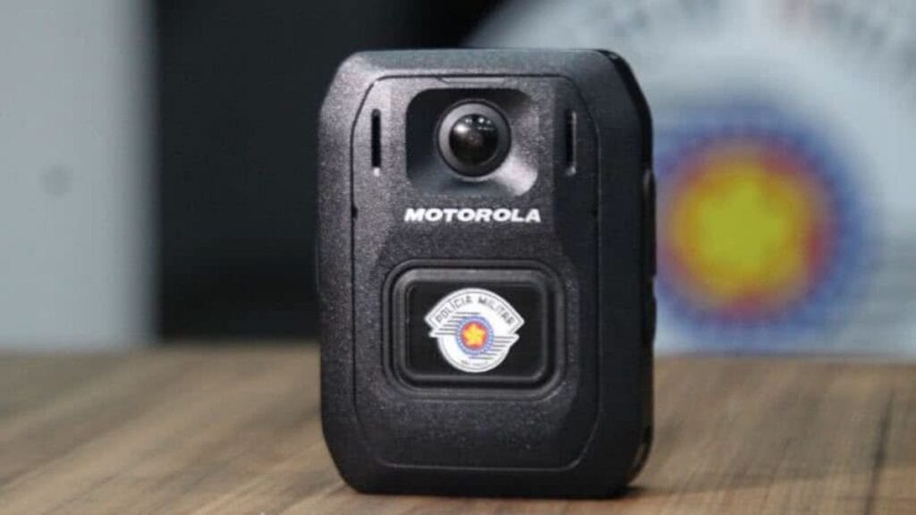 Câmera corporal da Motorola, utilizada pela Polícia Militar de São Paulo, aparece em destaque sobre uma superfície de madeira. O dispositivo exibe o brasão da corporação, simbolizando seu uso oficial. Ao fundo, desfocado, aparece outro brasão da PM paulista, reforçando o contexto institucional da imagem.