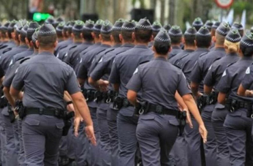 Altura mínima na PM vira questão de justiça: Na imagem vemos um grupo de policiais uniformizados marchando em formação, todos vestidos com fardas cinzas e boinas padronizadas, vistos de costas em um deslocamento coletivo.