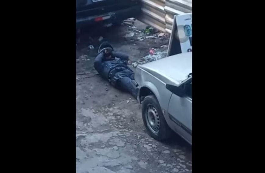 Policiais feridos: Imagem mostra um policial militar deitado no chão, com uniforme e capacete, falando no rádio, ao lado de um carro e próximo a um muro metálico.