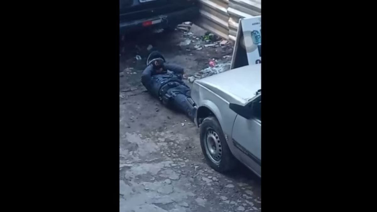 Policiais feridos: Imagem mostra um policial militar deitado no chão, com uniforme e capacete, falando no rádio, ao lado de um carro e próximo a um muro metálico.