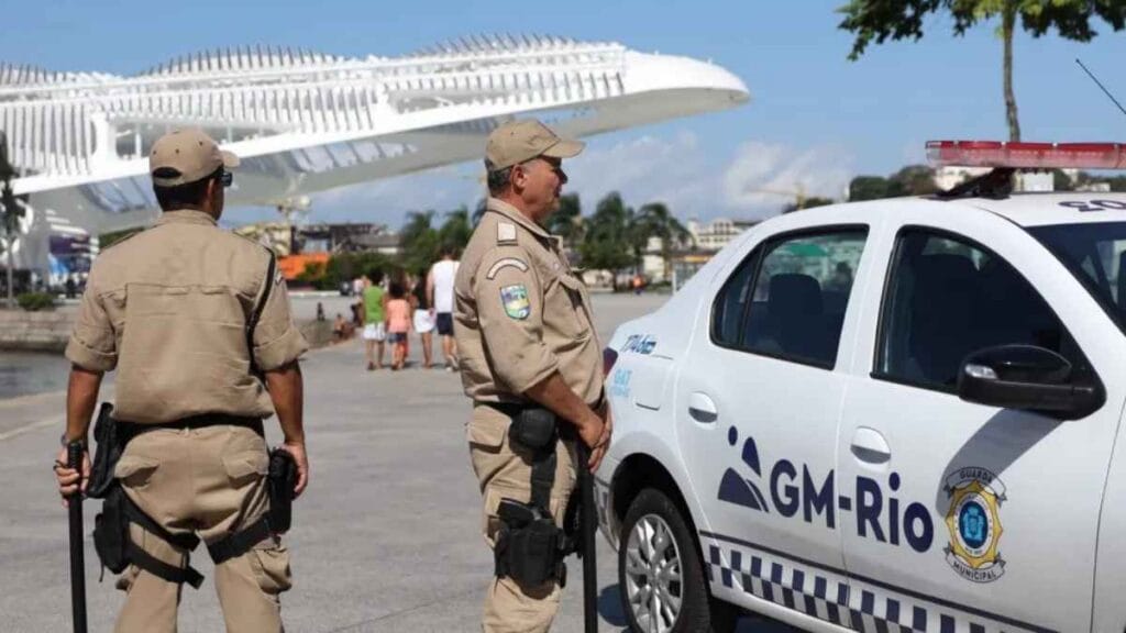 Força Municipal: Dois guardas municipais uniformizados de pé ao lado de uma viatura da GM-Rio, com o Museu do Amanhã ao fundo, em um dia ensolarado.