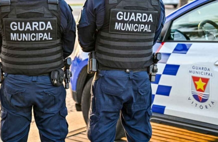 Estatuto Geral das Guardas Municipais: Dois agentes da Guarda Municipal de Vitória (ES), uniformizados e armados, estão de costas ao lado de uma viatura oficial com o brasão da corporação visível.