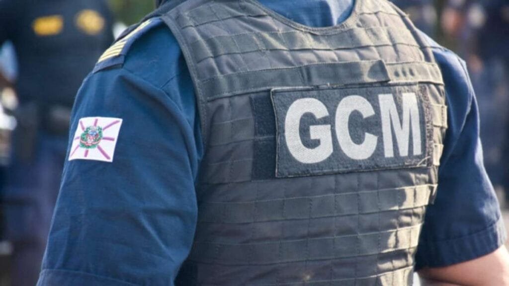 GCM: efetivo, desafios e o papel na segurança pública: Um guarda municipal aparece de uniforme azul e colete balístico preto, com a sigla **“GCM”** em destaque nas costas. No braço esquerdo, há um brasão com bandeira estilizada. A imagem foca no torso do agente, transmitindo a ideia de segurança e presença ostensiva.