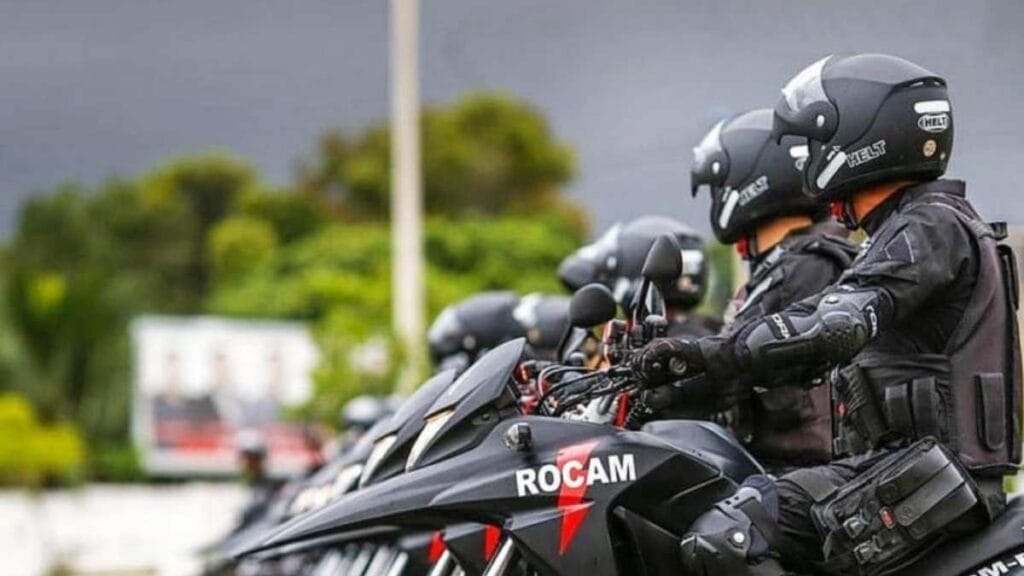 Policiais feridos: Imagem mostra policiais militares da ROCAM alinhados em motocicletas pretas, usando capacetes e equipamentos de proteção, prontos para operação.
