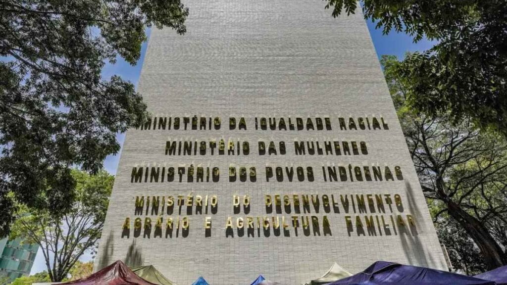 Alta violência contra mulher: Prédio em Brasília com fachada mostrando em destaque os nomes de quatro órgãos: **Ministério da Igualdade Racial, Ministério das Mulheres, Ministério dos Povos Indígenas e Ministério do Desenvolvimento Agrário e Agricultura Familiar**. Árvores aparecem ao redor e barracas coloridas estão montadas na parte inferior da imagem.
