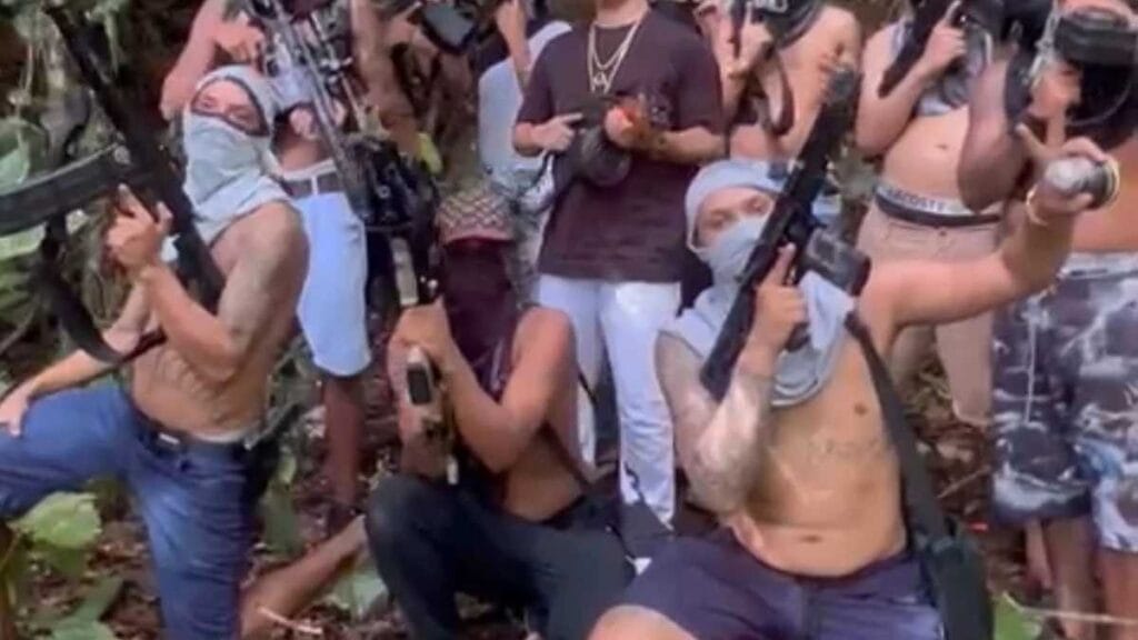 Domínio das facções: Grupo de homens armados, muitos com o rosto coberto por panos, exibindo fuzis e pistolas em área de mata, posando de forma ostensiva para a câmera.
