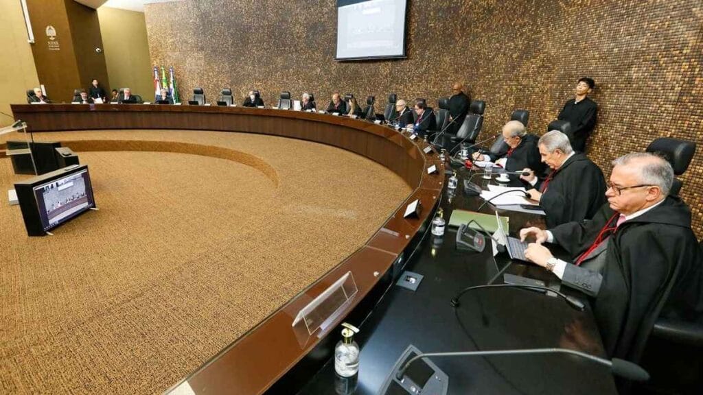 Sessão de julgamento no Tribunal de Justiça, com desembargadores e autoridades reunidos em mesa em formato semicircular. Alguns utilizam computadores e documentos durante a reunião.