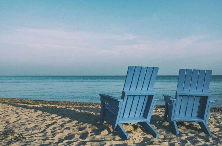 Aposentadoria Policial Duas cadeiras de madeira azuis posicionadas na areia de uma praia, voltadas para o mar calmo sob céu azul claro.