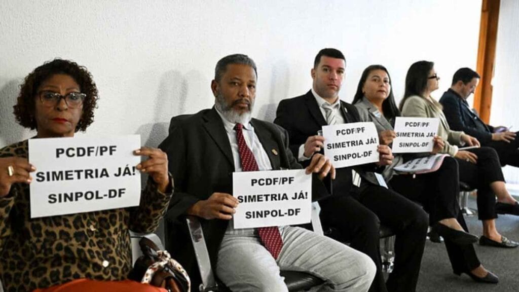 Grupo de pessoas sentadas em uma sala, segurando cartazes com a frase: “PCDF/PF SIMETRIA JÁ! SINPOL-DF”. Trata-se de uma manifestação pacífica organizada pelo sindicato da Polícia Civil do Distrito Federal, cobrando equiparação salarial com a Polícia Federal.