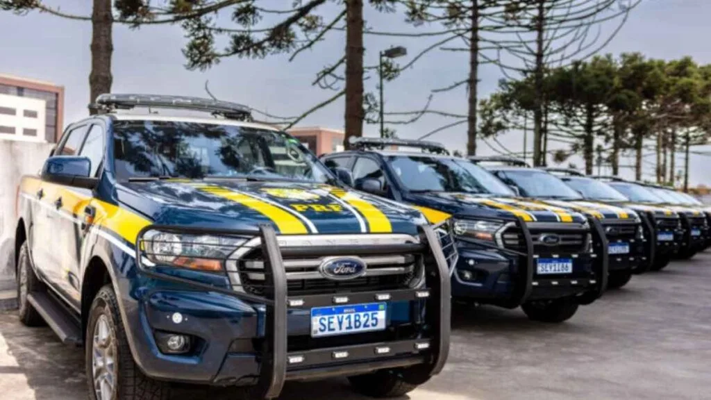 Fila de viaturas da Polícia Rodoviária Federal (PRF), modelo Ford Ranger caracterizadas nas cores azul e amarela da corporação, estacionadas em área externa, com árvores ao fundo.
