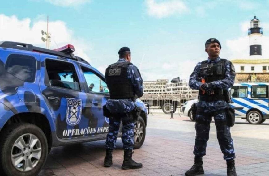 Os grupos especiais da GCM de Salvador: Dois agentes da Guarda Civil Municipal de Salvador, do Grupamento de Operações Especiais (GOE), estão posicionados em frente ao Farol da Barra. Eles usam fardamento tático azul camuflado, coletes e armamento. Ao lado, uma viatura oficial do GOE com pintura camuflada azul e a inscrição “Operações Especiais”.