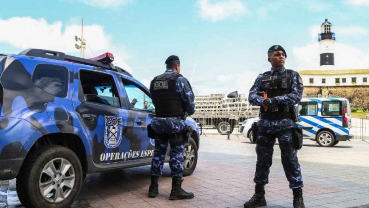 Os grupos especiais da GCM de Salvador: Dois agentes da Guarda Civil Municipal de Salvador, do Grupamento de Operações Especiais (GOE), estão posicionados em frente ao Farol da Barra. Eles usam fardamento tático azul camuflado, coletes e armamento. Ao lado, uma viatura oficial do GOE com pintura camuflada azul e a inscrição “Operações Especiais”.