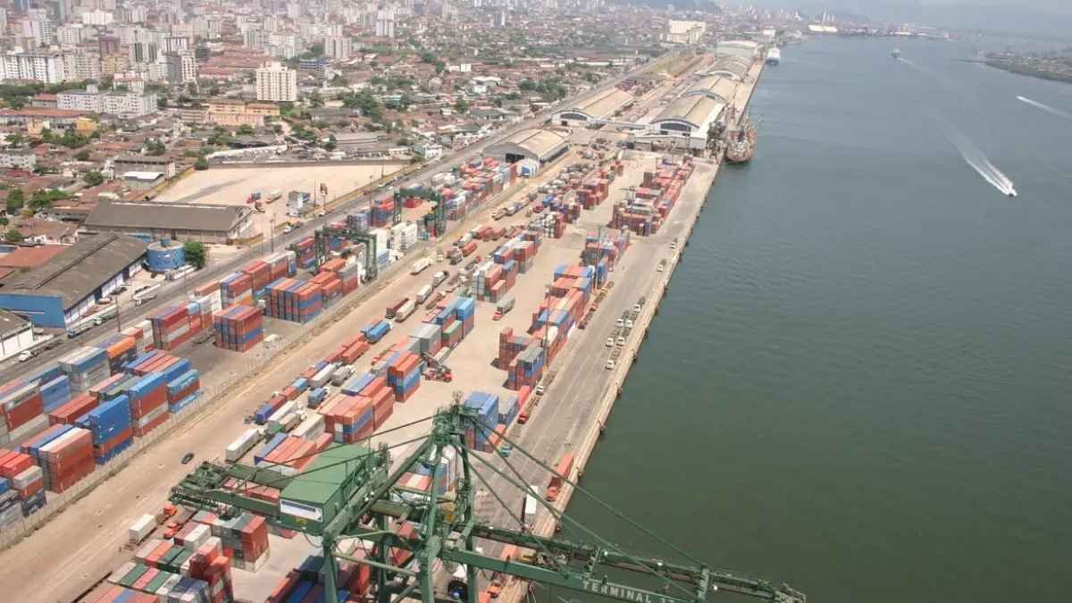 Brasil: entreposto de drogas. Porto com diversos contêineres coloridos organizados em pilhas, guindastes para carga e descarga e navios atracados, com vista da cidade ao fundo e o mar à direita.