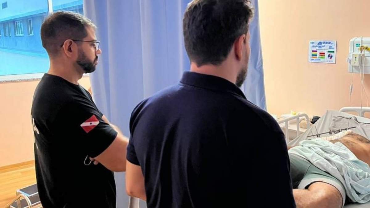 Policial penal assassinado durante custódia em Hospital: Dois homens observam paciente deitado em cama de hospital, parcialmente coberto por lençol.