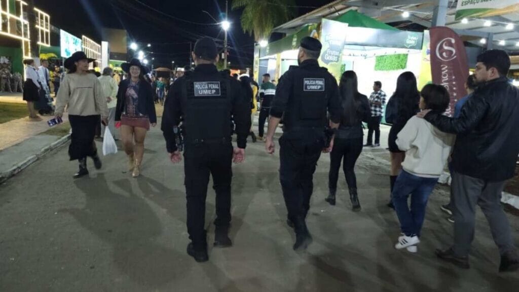 Agentes da Polícia Penal do Acre, identificados pelos uniformes com a sigla "UMEP", patrulham uma área pública durante evento noturno, cercados por pessoas caminhando em clima tranquilo. A cena mostra a presença da corporação reforçando a segurança em espaços urbanos frequentados por famílias.
