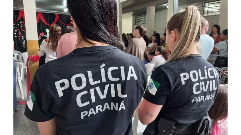 Envelhecimento das Tropas: Duas policiais civis do Paraná, de costas, vestindo camisetas pretas com a inscrição “POLÍCIA CIVIL PARANÁ”. Elas estão em um ambiente interno com outras pessoas ao fundo, em um evento coletivo.
