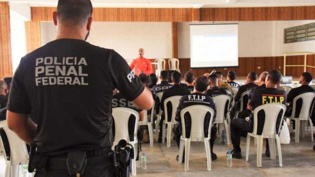questão de tablets: Policiais penais federais participam de capacitação em ambiente interno, sentados em cadeiras brancas de plástico, enquanto acompanham uma apresentação projetada em telão. Em destaque, um agente de costas com o uniforme da Polícia Penal Federal. A formação reúne integrantes de diferentes unidades especializadas, como o grupo F.T.I.P, reforçando a qualificação contínua da corporação.