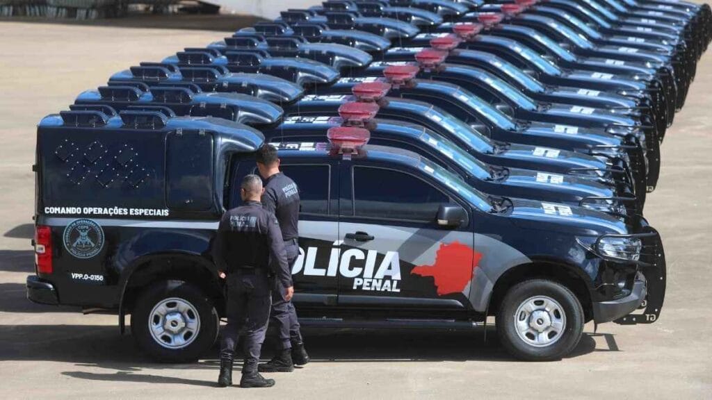 Dois policiais penais observam uma frota de viaturas novas da Polícia Penal, enfileiradas em pátio aberto, identificadas com logotipo institucional e luzes de emergência no teto.
