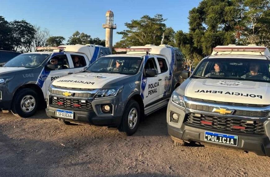 Viaturas da Polícia Penal do Paraná estão alinhadas em uma área aberta durante o dia, com agentes no interior dos veículos prontos para operação. As camionetes caracterizadas evidenciam a estrutura e presença da corporação no policiamento externo e no transporte de presos.