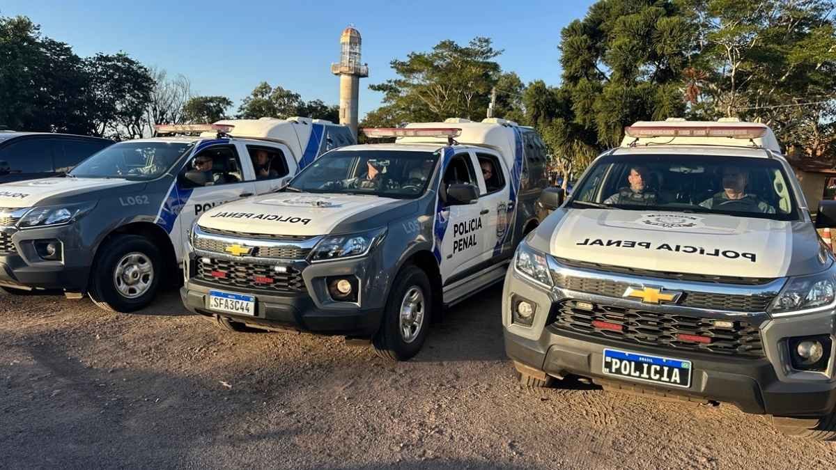 Viaturas da Polícia Penal do Paraná estão alinhadas em uma área aberta durante o dia, com agentes no interior dos veículos prontos para operação. As camionetes caracterizadas evidenciam a estrutura e presença da corporação no policiamento externo e no transporte de presos.