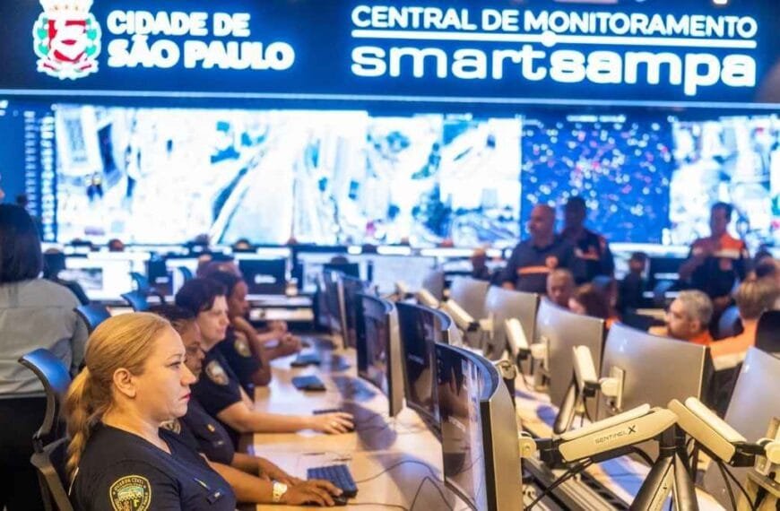 Grande irmão: Sala de monitoramento da Prefeitura de São Paulo, com agentes da GCM e técnicos acompanhando imagens em tempo real em vários monitores; ao fundo, painel eletrônico com a inscrição “Cidade de São Paulo – Central de Monitoramento Smart Sampa”.