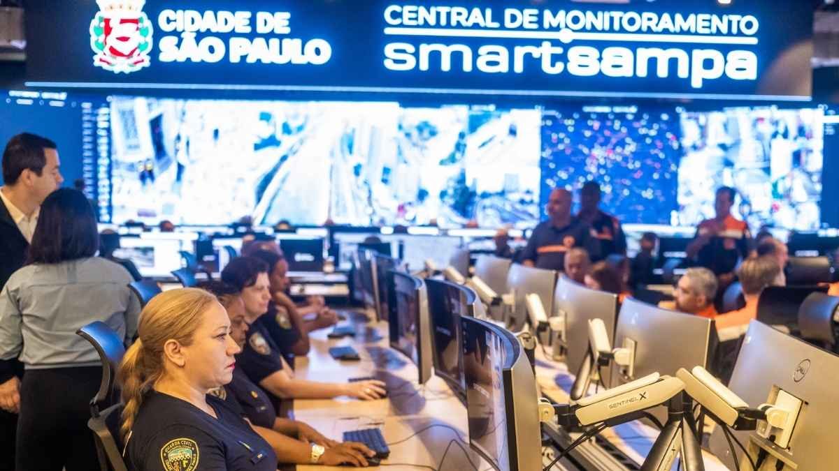 Grande irmão: Sala de monitoramento da Prefeitura de São Paulo, com agentes da GCM e técnicos acompanhando imagens em tempo real em vários monitores; ao fundo, painel eletrônico com a inscrição “Cidade de São Paulo – Central de Monitoramento Smart Sampa”.