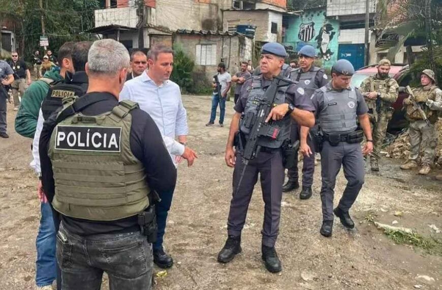 Mortes de policiais crescem 30%: Grupo de policiais militares e civis armados, acompanhados de autoridades, reunidos em uma comunidade com casas simples ao fundo.