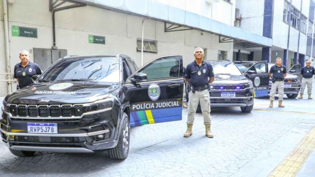 Cinco agentes da Polícia Judicial posam ao lado de viaturas pretas modelo Jeep, estacionadas em frente a um prédio do Tribunal Regional do Trabalho (TRT). Os veículos exibem o emblema da Justiça do Trabalho e a inscrição "Polícia Judicial". O local possui sinalizações nas paredes com indicações de blocos. A cena transmite organização e presença institucional.

