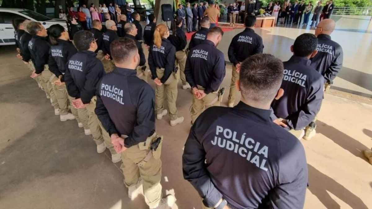 Polícia Judicial: Grupo de policiais judiciais, uniformizados com calças caqui e blusas pretas com a inscrição "Polícia Judicial", está perfilado de costas em posição de respeito durante cerimônia oficial. À frente, autoridades discursam em um palanque, enquanto convidados acompanham o evento. A cena destaca a organização, disciplina e formalidade da corporação em contexto institucional.