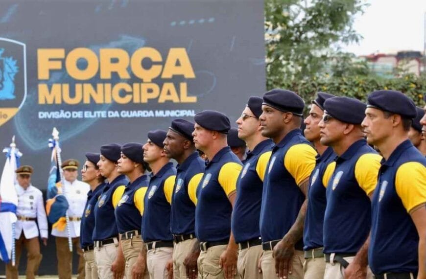 Integrantes da Guarda Municipal do Rio, uniformizados com boinas e camisas azul e amarela, estão perfilados em formatura. Ao fundo, um painel exibe a inscrição “Força Municipal – Divisão de Elite da Guarda Municipal”.