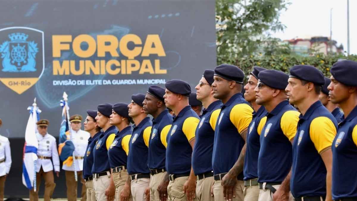 Integrantes da Guarda Municipal do Rio, uniformizados com boinas e camisas azul e amarela, estão perfilados em formatura. Ao fundo, um painel exibe a inscrição “Força Municipal – Divisão de Elite da Guarda Municipal”.