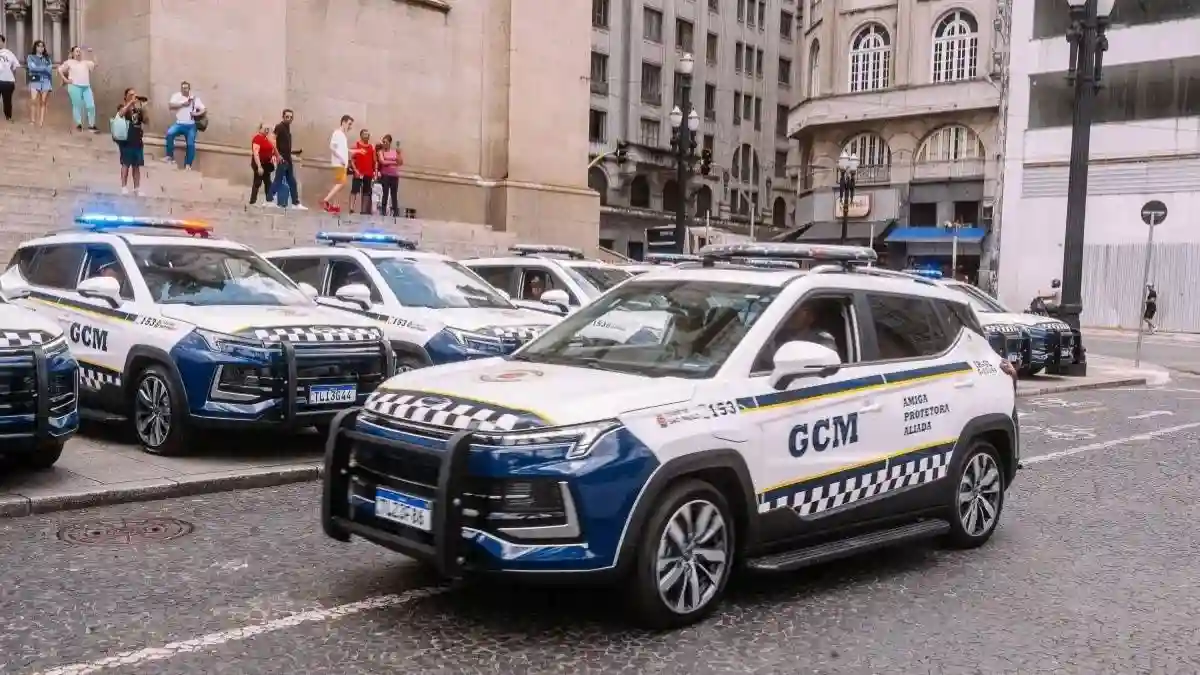 GCM de São Paulo e São Caetano atuam juntas - A imagem mostra viaturas da **Guarda Civil Metropolitana (GCM) de São Paulo**, alinhadas em via pública no centro da cidade, com sirenes ligadas. Os veículos têm pintura branca e azul, com faixas quadriculadas e o brasão da corporação. Ao fundo, pessoas observam próximas a uma escadaria de prédio histórico.