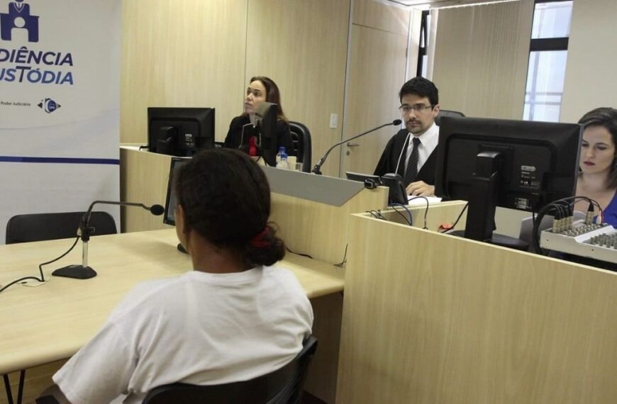 Juíza quase leva preso pra casa-Texto alternativo resumido: Pessoa participa de uma audiência de custódia em sala de tribunal; à frente dela estão juíza, servidores e equipamentos de informática. Um banner com o texto “Audiência de Custódia” está visível ao lado.