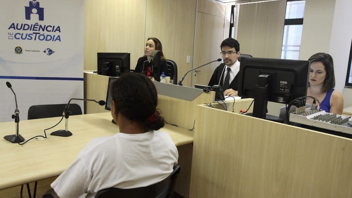 Juíza quase leva preso pra casa-Texto alternativo resumido: Pessoa participa de uma audiência de custódia em sala de tribunal; à frente dela estão juíza, servidores e equipamentos de informática. Um banner com o texto “Audiência de Custódia” está visível ao lado.