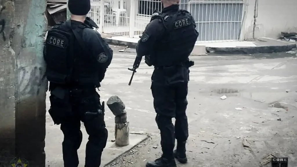 Dois agentes fortemente armados, com uniformes táticos da CORE (unidade de operações especiais da polícia), estão de costas em uma rua urbana, durante uma ação em área residencial.