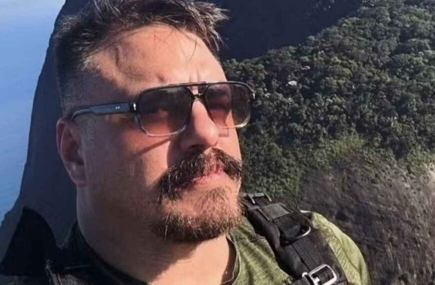 Delegado da PF preso! Facilitou tráfico de armas e drogas. Homem de óculos escuros e bigode aparece em primeiro plano, vestindo camiseta verde e mochila. Ao fundo, vê-se uma paisagem com vegetação e encosta íngreme próxima ao mar.