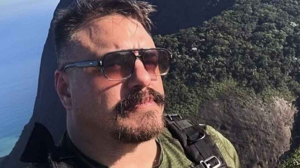 Delegado da PF preso! Facilitou tráfico de armas e drogas. Homem de óculos escuros e bigode aparece em primeiro plano, vestindo camiseta verde e mochila. Ao fundo, vê-se uma paisagem com vegetação e encosta íngreme próxima ao mar.