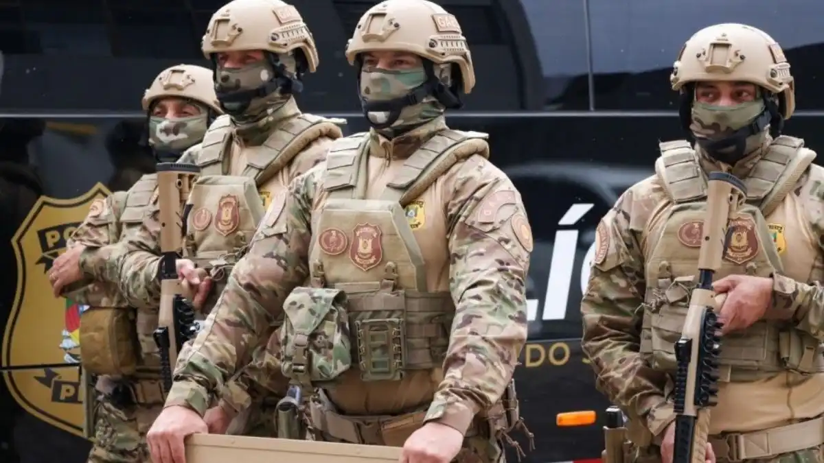 Novo uniforme: PPRS -Homens fardados da Polícia Penal do Rio Grande do Sul, integrantes do Grupo de Ações Especiais (GAES), posam lado a lado usando novos uniformes camuflados em tons de bege e verde, coletes balísticos e capacetes, com armas e equipamentos táticos diante de viatura oficial.