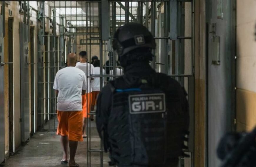 Presídio Central de Porto Alegre-Agente da Polícia Penal observa detentos uniformizados andando em corredor de cela dentro de unidade prisional