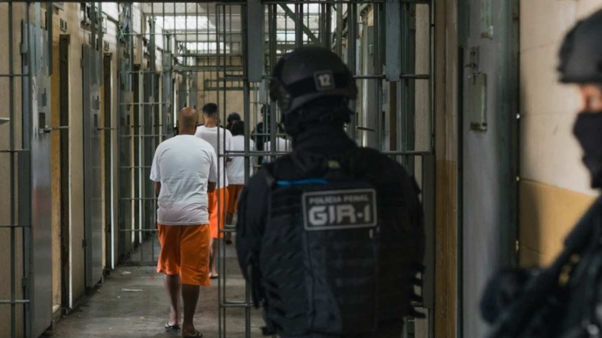 Presídio Central de Porto Alegre-Agente da Polícia Penal observa detentos uniformizados andando em corredor de cela dentro de unidade prisional