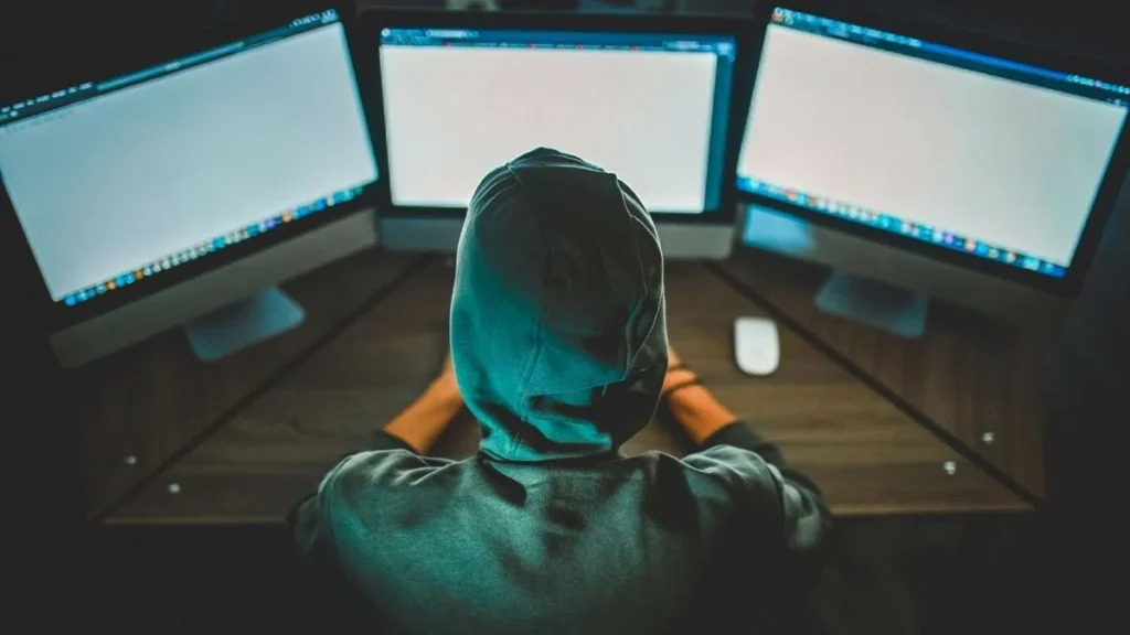 Uma pessoa de capuz, de costas, sentada em uma mesa e usando três monitores de computador.