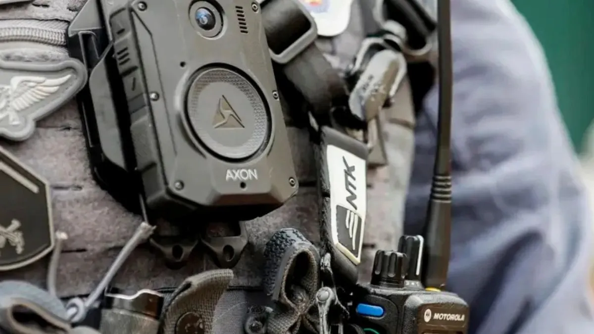 A imagem mostra de perto o colete de um policial equipado com **câmera corporal (bodycam) da marca Axon**, fixada na parte frontal do uniforme. Também é possível ver rádios comunicadores e outros acessórios táticos utilizados em operações. Governo Federal foca em câmeras para GCM's
