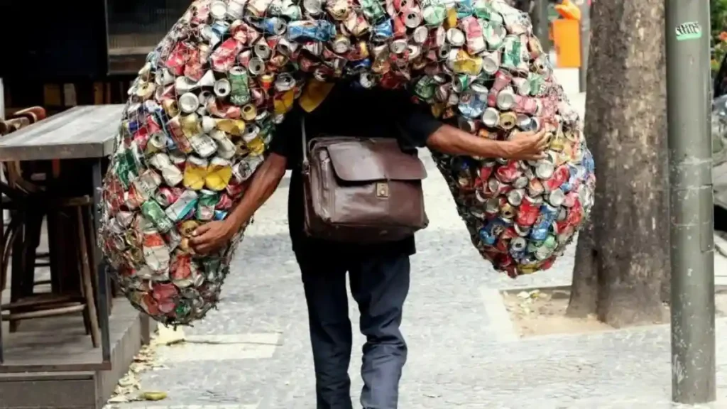 A imagem mostra um homem carregando nas costas um grande volume de latas de alumínio amassadas, presas em uma rede. Ele caminha por uma calçada e usa uma bolsa de couro marrom a tiracolo.
