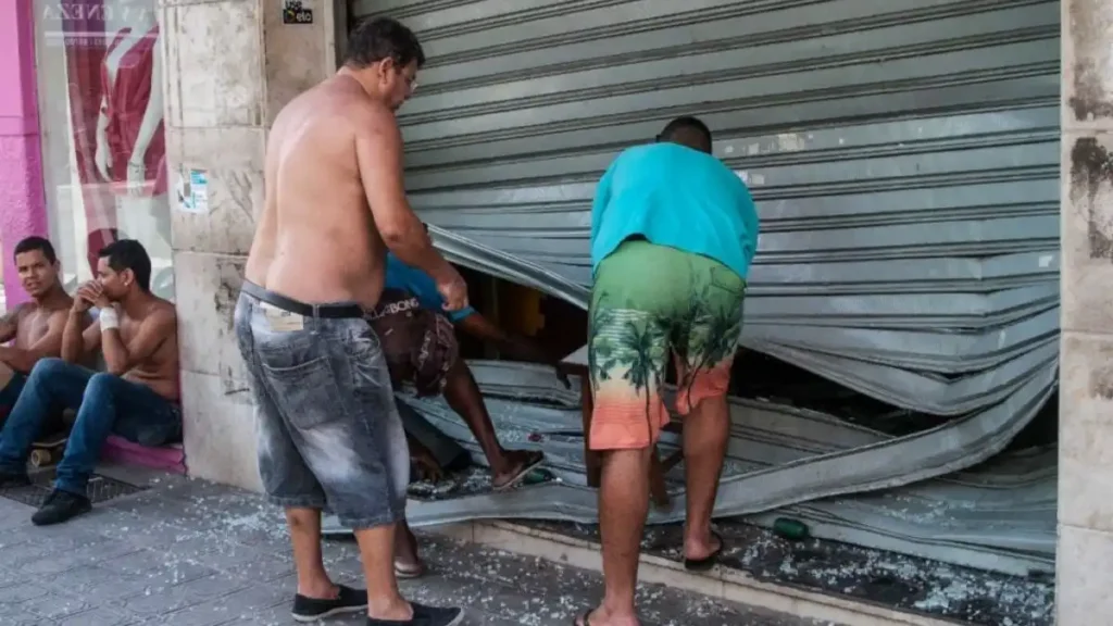 Homens levantam parcialmente a porta de aço de uma loja danificada para entrar no interior, em meio a estilhaços de vidro espalhados pelo chão.
