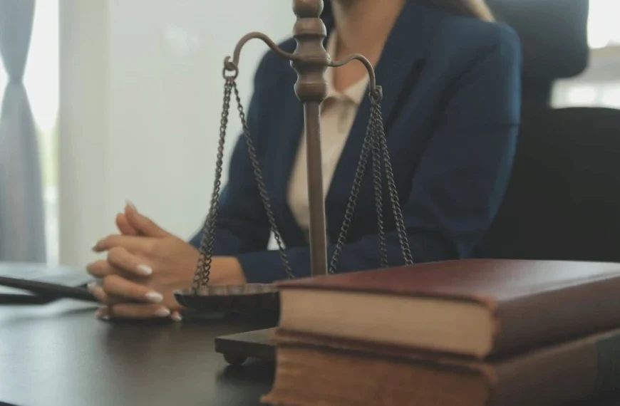 Falso juiz-A imagem mostra uma mulher sentada atrás de uma mesa de escritório, vestindo blazer azul. Em primeiro plano aparecem uma balança da Justiça e dois livros empilhados.