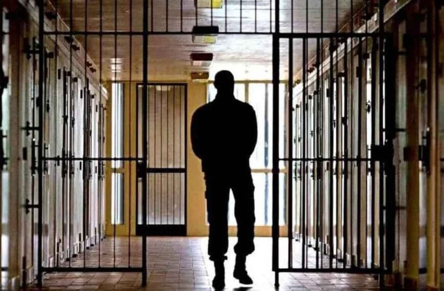 Matou e concretou a mãe; foi solto e...Homem em silhueta caminha por corredor de presídio, cercado por celas gradeadas e iluminação natural ao fundo.