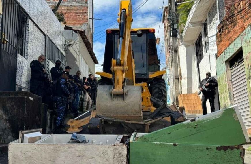 **Texto alternativo resumido da imagem:** Uma retroescavadeira remove barricadas feitas com geladeiras e entulhos em uma rua estreita de Salvador, enquanto vários policiais armados observam e dão apoio à operação.